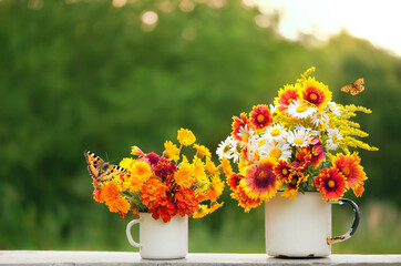 two enamel mugs with colorful flowers and butterflies on table in garden, natural background. summer season. rustic composition with flowers. relax time.