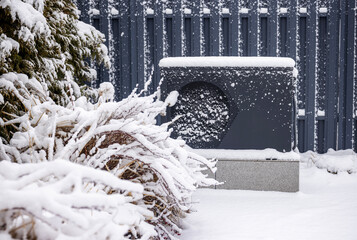 Snow-covered outdoor heat pump unit beside frosted bushes and conifers against a gray metal fence...