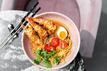 A bowl of ramen with crispy fried shrimp, halved boiled egg, cherry tomatoes, and cilantro, placed on a textured surface with chopsticks. The dish is vibrant, fresh, and appetizing