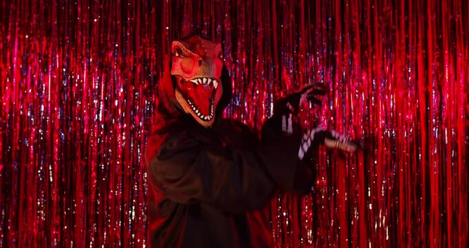 Spooky dinosaur in black cloak moving and gesturing actively trying to frighten you, being on Halloween party, posing against red shiny background. Unknown guy using animal style on masquerade