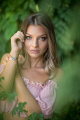 Naklejka premium portrait of a young beautiful woman with striking green eyes,captured in a natural outdoor setting.She is framed by lush green foliage. Woman dressed in a light pink off-shoulder top and skirt posing
