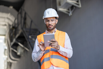 An engineer uses a tablet, wearing a safety vest, glasses, and hard hat. Indoors setting.