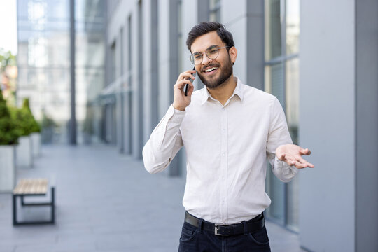 Fototapeta A smiling businessman talks on his phone in front of a modern building. He gestures with his hand while discussing something.