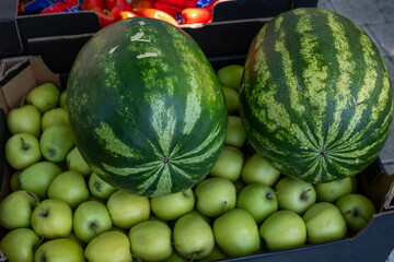 Two large, striped watermelons rest on top of a generous crate of bright green apples, creating an abundant display of fresh, ripe summer fruits at a market stall