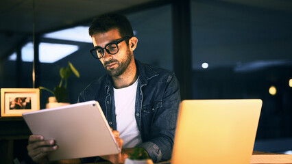 Tablet, laptop and man in office at night with creative project with publishing online. Glasses,...
