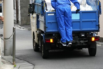 a Japanese garbage or utility truck, with a worker in blue overalls standing on the back, appearing to oversee the loaded materials or preparing for collection.A narrow urban street or alleyway,city l