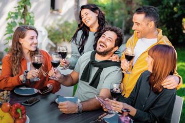 Happy group of young friends laughing and drinking red wine at outdoor garden party in summer.