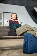 Relaxed Eastern European woman talking on phone at bus stop with suitcase