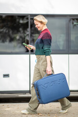 Cheerful female traveler with short blond hair checking phone while walking past bus