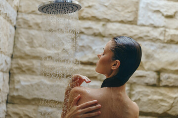 woman showering under rainfall showerhead with stone wall background, wet hair, relaxed, natural skincare, spa atmosphere, wellness concept