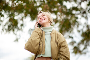 Cheerful blond mature woman talking on phone outdoors under leafy autumn trees