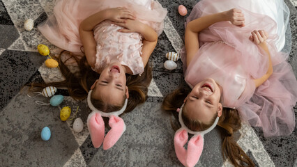 Cute laughing little girls with bunny ears are lying on a floor with colorful eggs in the Easter holiday.