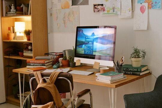 Home workspace featuring modern desktop computer on wooden desk surrounded by books, potted plants, school supplies, backpack and children's drawings on wall emphasizing family environment