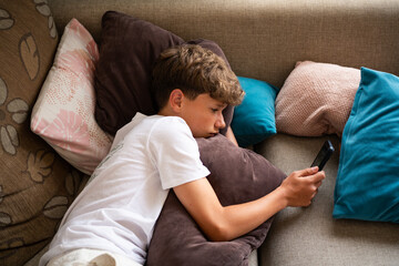 A young boy lies comfortably on a couch, resting his head on soft pillows. He focuses intently on...