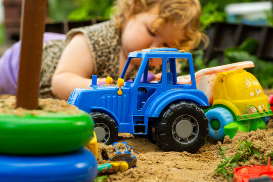 Blue toy tractor moving through sand with children playing nearby at a sunny outdoor location