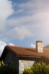 A rustic stone house roof with terracotta tiles and a stone chimney, framed by pine tree branches and set against a clear blue sky.