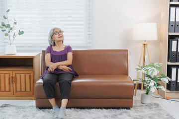 Retired Asian woman sitting comfortably on a sofa in her living room, gazing thoughtfully out the...