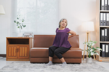 Elderly asian woman experiencing back pain while sitting on a sofa in her living room, demonstrating the common issue of discomfort and mobility challenges faced by seniors at home
