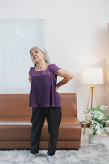Elderly asian woman suffering from backache, standing in living room, holding hands on painful...