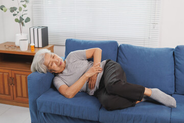 Elderly Asian woman lying on a sofa, clutching her stomach in distress, experiencing abdominal...