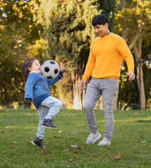 Happy latin dad and son kicking soccer ball in park, enjoying leisure time together on sunny autumn...