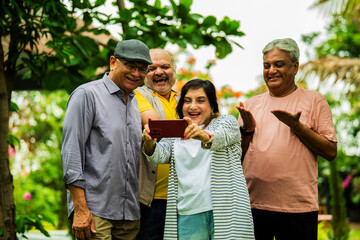 Checking photos on smartphone, elderly Indian group smiles while standing in lush garden