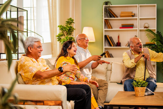 Indian seniors watching television laughing together on sofa during joyful home reunion gathering