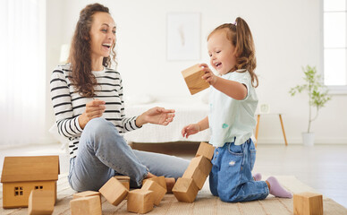 Happy cheerful family laughing after destroying block toy tower. Joyful mother playing educational game with wooden cubes and stacking blocks with her small preschool daughter in spacious bedroom.