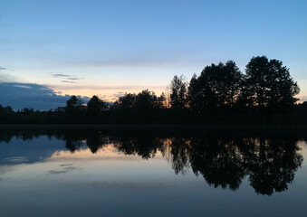Beautiful twilight sunset. The dark dense summer forest is reflected in the mirrored lake. Bright...