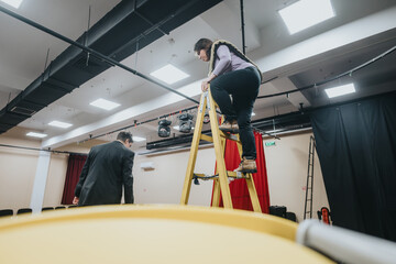 Two individuals, one climbing a ladder, prepare props in a theatrical setting under rehearsed...