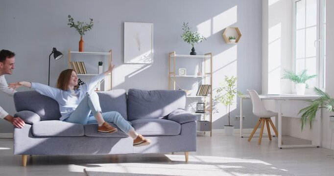 Smiling husband playfully pushing sofa with wife sitting on it in their new home. Joyful couple hugging on couch, feeling excited and celebrating successful move and beginning of new chapter together.