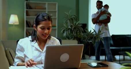 Indian mother celebrates success working on laptop while managing documents and phone call at home, professional woman joyful as father walks behind with baby boy in modern Indian family setting - Powered by Adobe