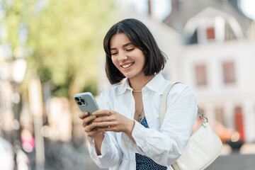 Fototapeta premium Street break. Indian woman in a white shirt with short hair types messages on her phone while commuting through the city with a soft smile