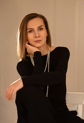 Elegant woman in black outfit and pearl necklace sitting on a chair indoors. Soft warm light, minimalist setting, confident facial expression. Studio portrait