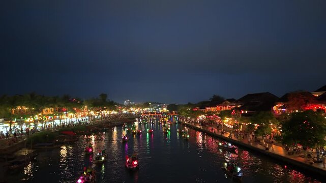 Drone flight over the Thu Bon River in the beautiful Vietnamese city of Hoi An at night. shows small boats on the water, iconic yellow buildings, colorful lanterns, and many people walking.