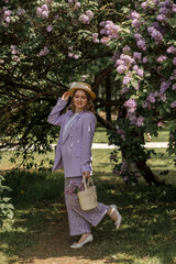 A beautiful young blonde woman, against a background of blooming lilacs, in a lilac trouser suit. Femininity and nature.
