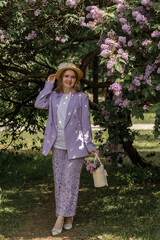 A beautiful young blonde woman, against a background of blooming lilacs, in a lilac trouser suit. Femininity and nature.