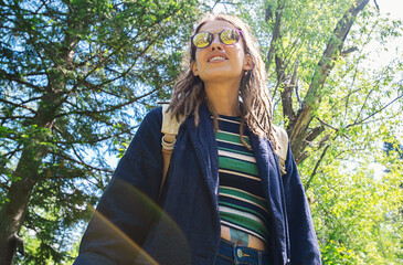Tourist Woman Stands in a Sunny Forest. She Wears a Striped Shirt and a Backpack, Surrounded by Green Trees.