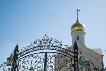 white brick Orthodox church with a golden dome and a cross against a clear blue sky, with a wrought-iron gate in the foreground
