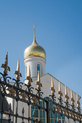 white brick Orthodox church with a golden dome and a cross against a clear blue sky, with a wrought-iron fence with pointed spires in the foreground