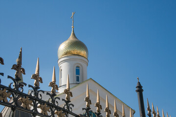 white brick Orthodox church with a golden dome and a cross against a clear blue sky, with a wrought-iron fence with pointed spires in the foreground