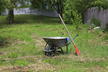 a metal garden wheelbarrow is in the garden, a shovel, a hose and blue work gloves are in it and...