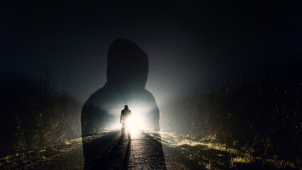 A double exposure of a hooded figure, with a mysterious figure silhouetted by spooky glowing lights on a country road at night