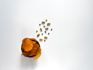 Unhealthy snack food and supplements. A bowl of potato chips next to a scattering of various pills and capsules on a white background. Choosing unhealthy eating over a healthy lifestyle.