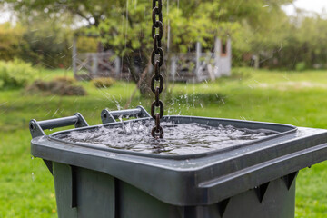 Rainwater chain splashes into plastic barrel in backyard garden, eco water harvesting solution for sustainable lifestyle, climate adaptation, rural off-grid living, green environment concept
