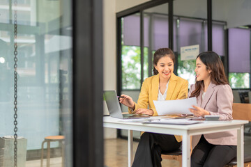 Two professional businesswomen are working on laptop computers sitting at desks in the office. Colleagues are happy and discussing the digital strategy of the Graph