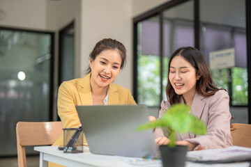 Two professional businesswomen are working on laptop computers sitting at desks in the office. Colleagues are happy and discussing the digital strategy of the Graph