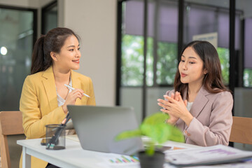 Two professional businesswomen are working on laptop computers sitting at desks in the office. Colleagues are happy and discussing the digital strategy of the Graph