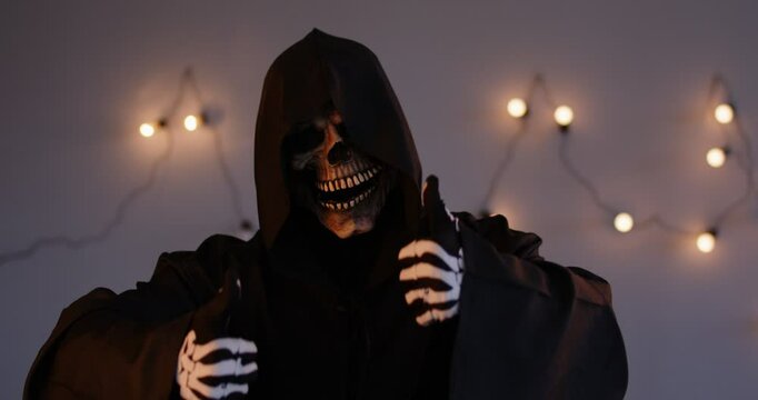 Portrait of person disguised in skull mask and skeleton grim reaper costume of death wearing black robe with hood, looking at camera and gesturing thumbs up on illuminated wall background. 