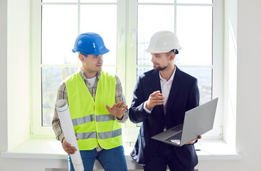 Portrait of young foreman discussing with professional architect design project or repair of new apartment on a laptop standing in empty room. Meeting with builder about interior decoration.